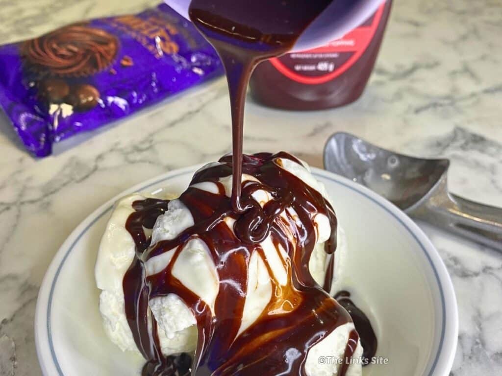 Vanilla ice cream in a white bowl covered in glossy hot fudge sauce, with a thin ribbon of sauce drizzling down from above in a square crop.