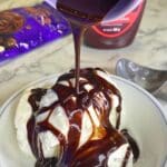 Vanilla ice cream in a white bowl covered in glossy hot fudge sauce, with a thin ribbon of sauce drizzling down from above in a square crop.